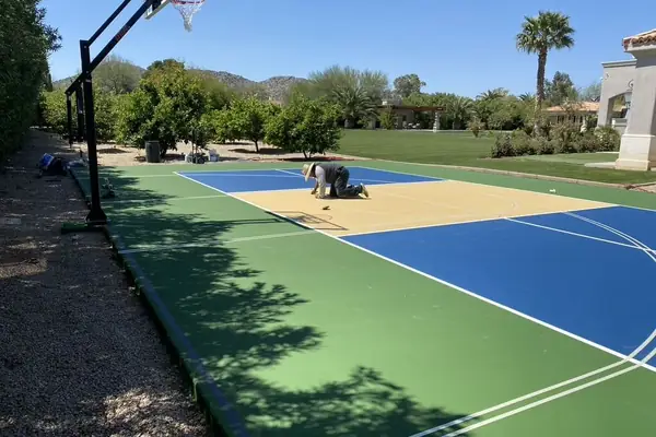 Crew member kneeling on a green, blue and tan court taping lines, hoop in the foreground