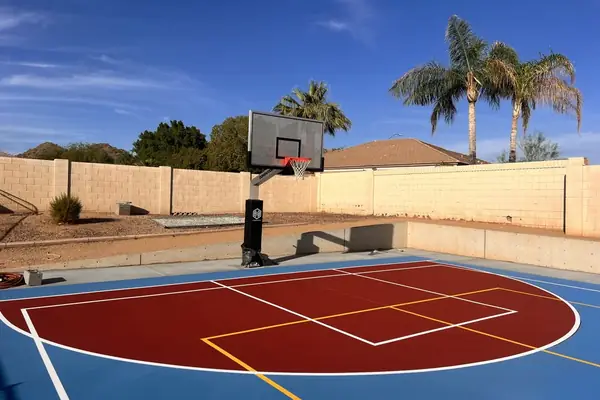 Backyard half court with a dark red key, yellow second lines and an in-ground hoop in front of a block wall and palms