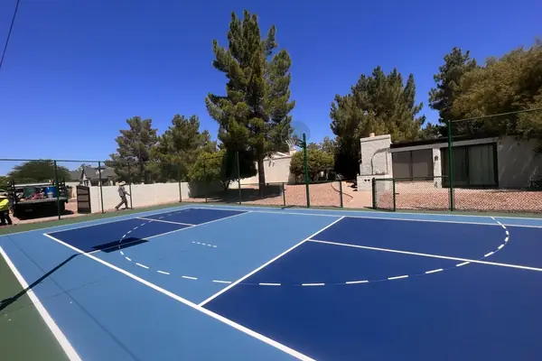 Two-tone blue court with pickleball and basketball lines, green fence and pine trees