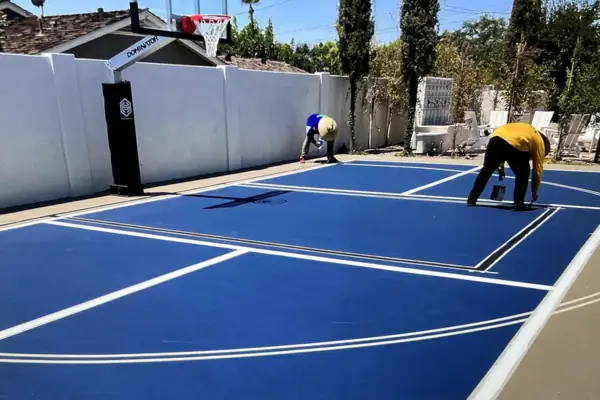 Two crew members coating a blue pickleball court beside a white block wall