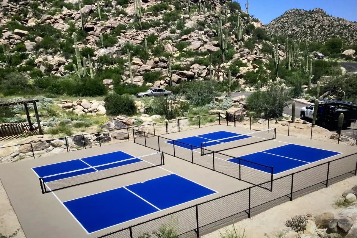 Four blue pickleball courts with black fencing on a desert hillside with saguaros