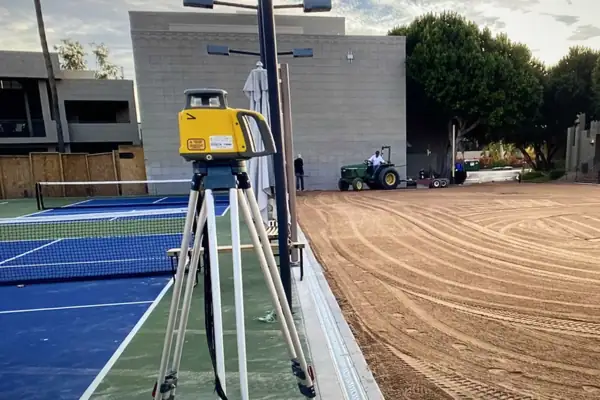 Laser level on a tripod over a graded pad beside a finished blue court