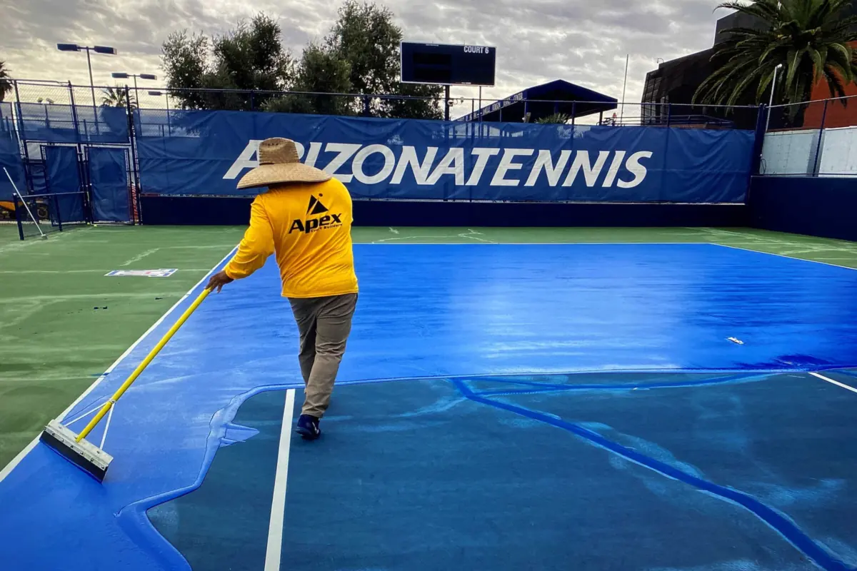 Apex crew member in a yellow Apex shirt pulling a squeegee of fresh blue coating across a tennis court