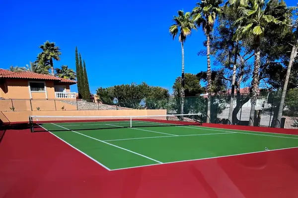 Red and green residential tennis court with a net, palms and a tile-roof house behind