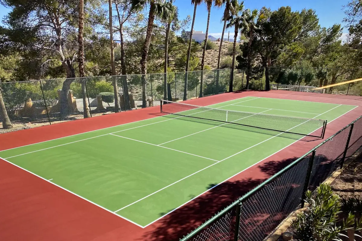 Red and green residential tennis court with a black chain link fence, palms and desert hills behind