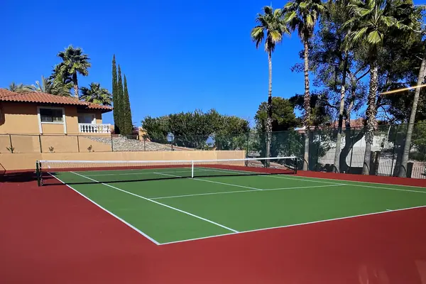 Red and green tennis court with a net, tile roofed house and palms behind