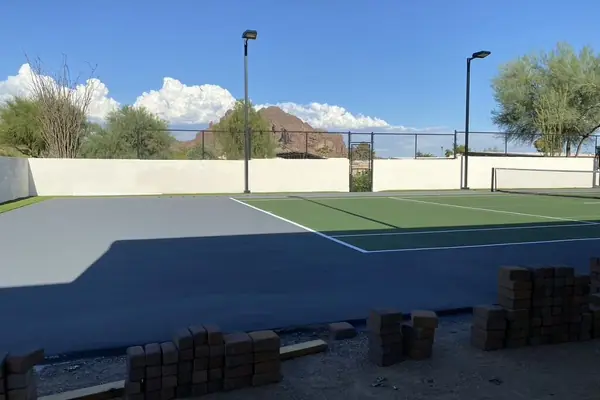 Fresh grey court pad beside a green tennis court with light poles, pavers stacked in front